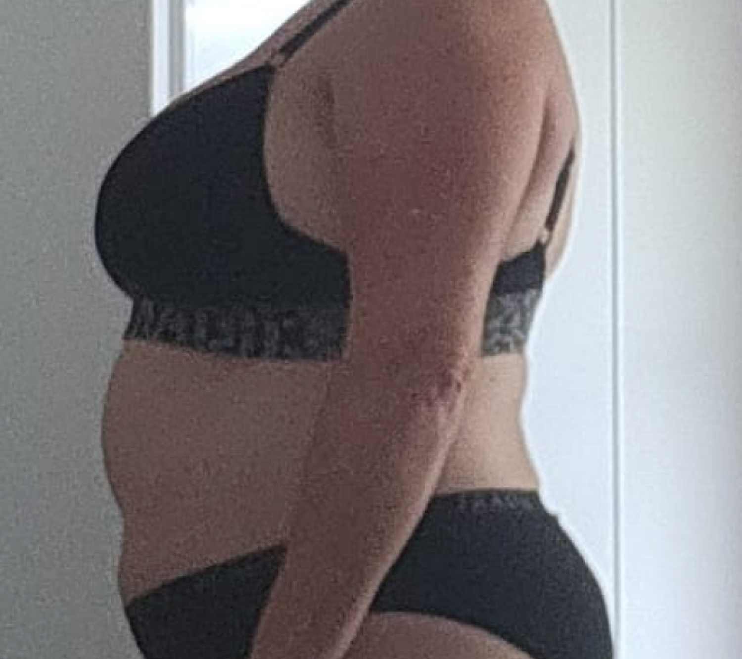 A person in black underwear stands sideways, showing their profile. The background is a softly lit interior.