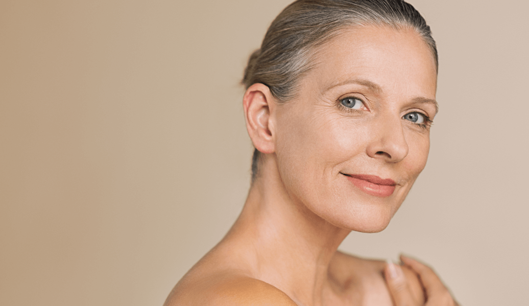 Middle-aged woman with smooth skin and subtle makeup, smiling at the camera. Background is a soft beige. Beauty and skincare concept.