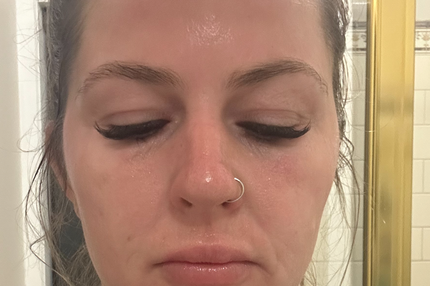 Woman with closed eyes, subtle makeup, and nose ring, standing in a bathroom. Focus on skincare, possibly post-collagen treatment.