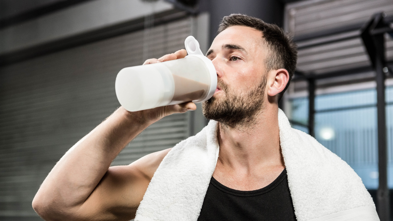 Man drinking collagen supplement shake in gym, wearing a towel and black tank top, promoting post-workout beauty and wellness.