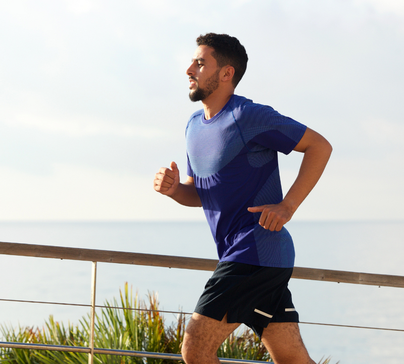 Man jogging by the sea, wearing a blue shirt and black shorts, railing and plants in the background, showcasing active lifestyle.