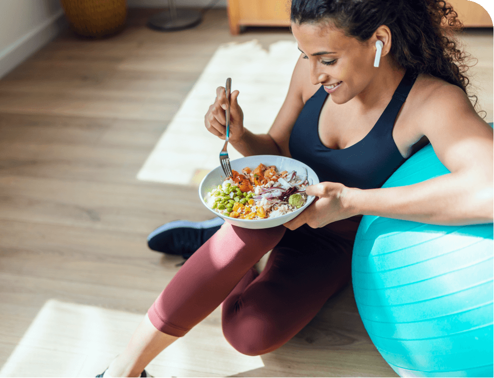 Woman in workout attire eating a healthy salad while sitting on an exercise ball, wearing wireless earbuds. Bright, airy room.
