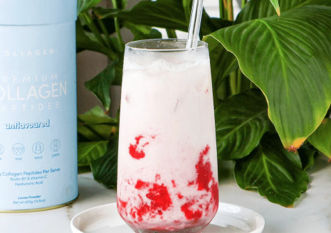Chilled collagen drink in a clear glass with red fruit swirls, beside a blue collagen peptide container, set against lush green leaves.