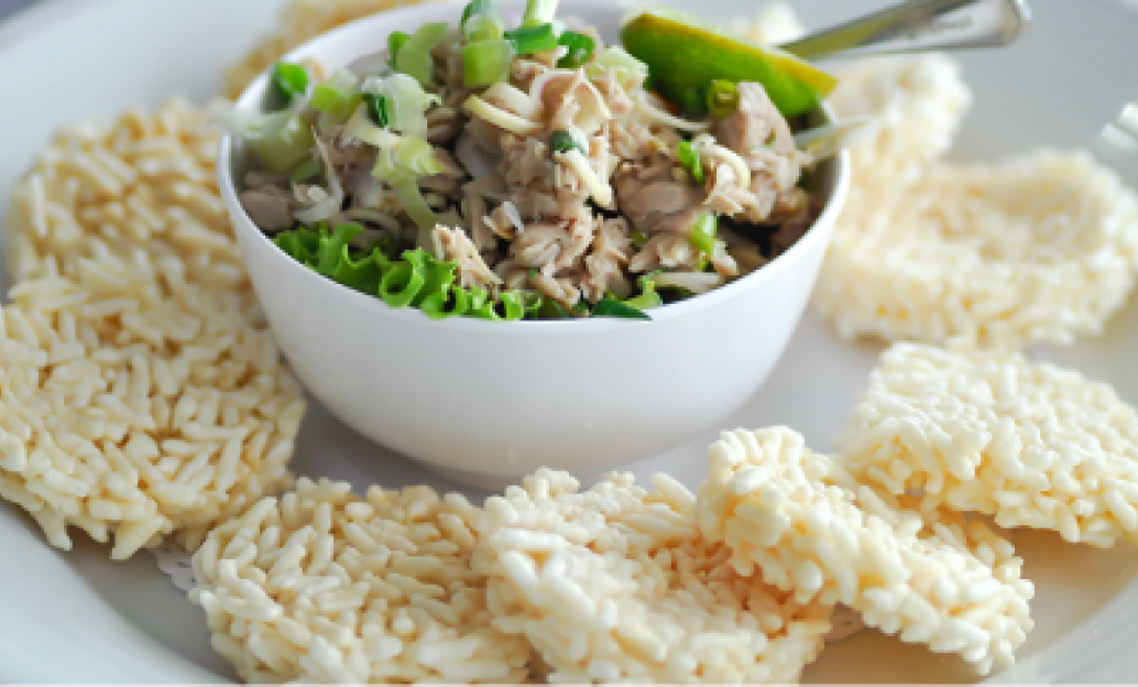White bowl with shredded chicken, green onions, and lime wedge, surrounded by crispy rice cakes on a white plate.