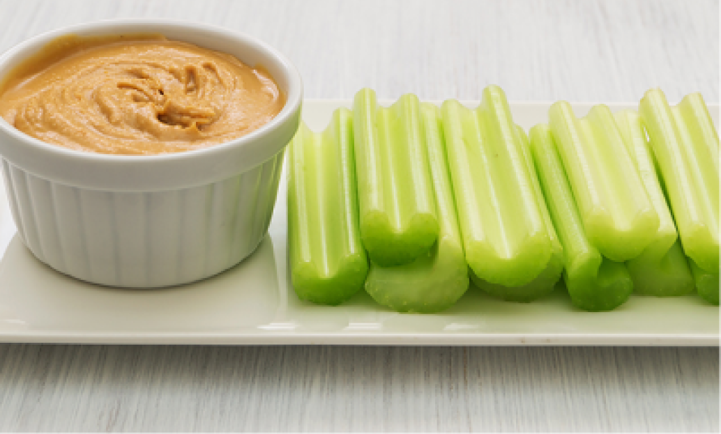 Plate with celery sticks and a small white dish of creamy peanut butter on a light wooden table. Perfect healthy snack option.