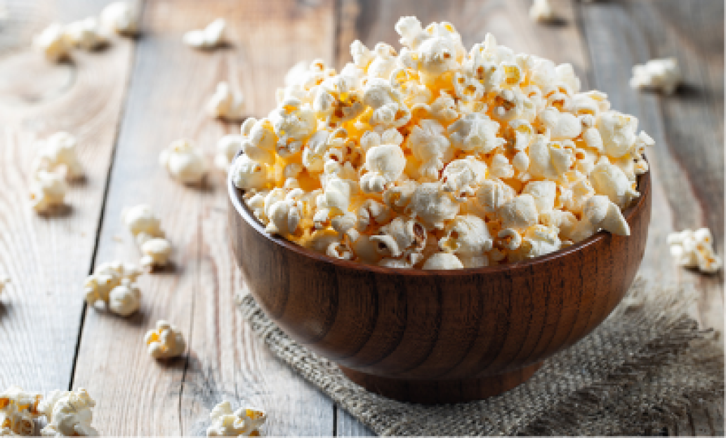 A wooden bowl filled with fluffy, freshly popped popcorn sits on a rustic wooden table, with scattered kernels around it.
