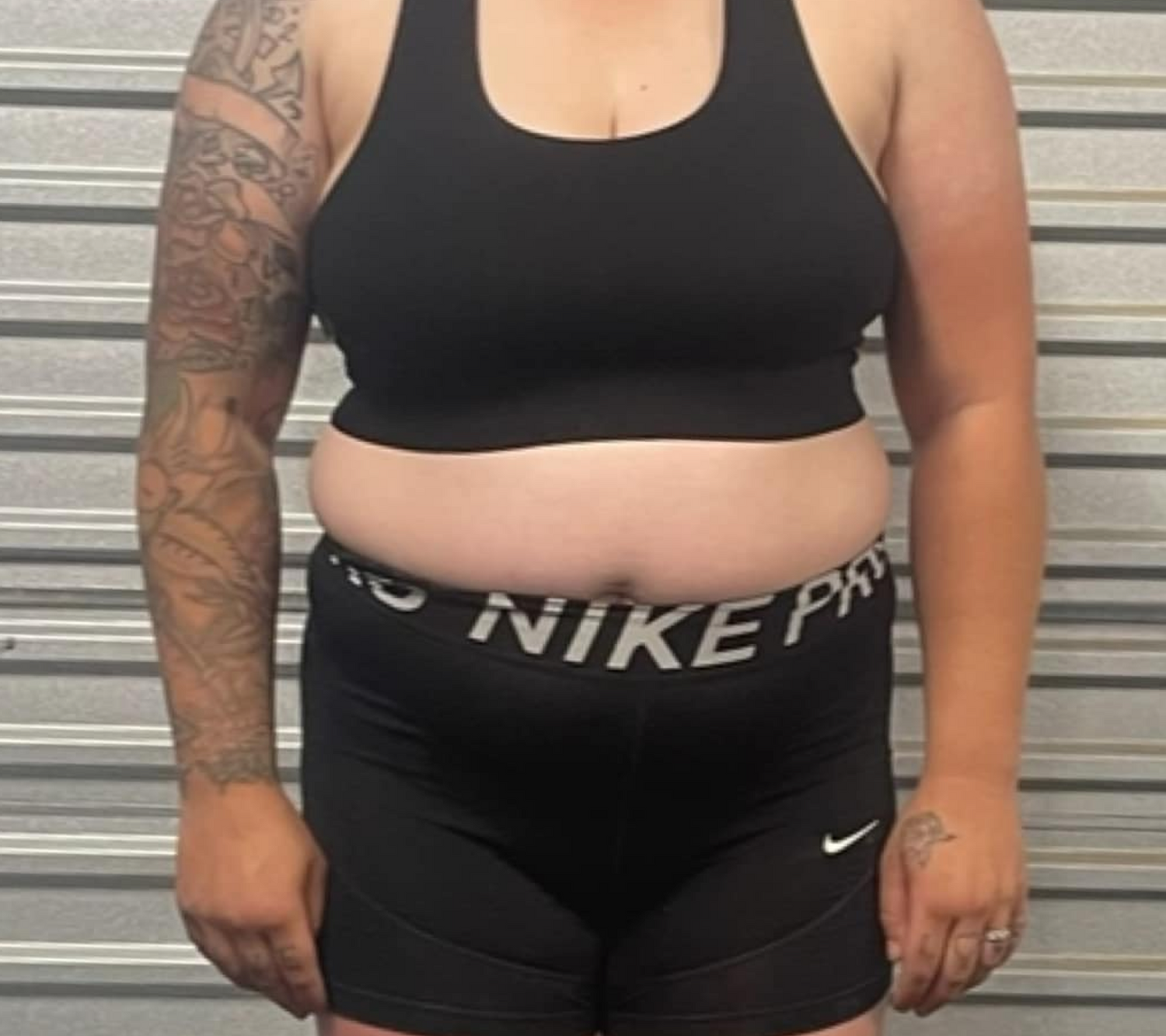 Person in black sportswear, showing tattooed arm and visible midsection, standing before a gray metal backdrop.