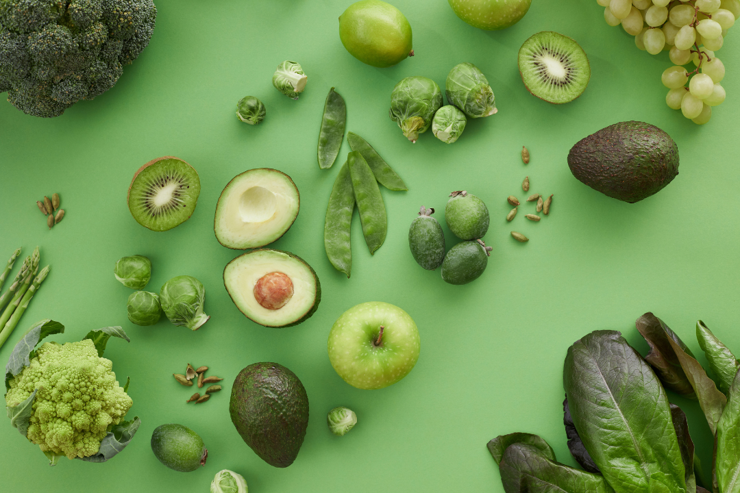 Assorted green fruits and vegetables on a green background, including avocado, kiwi, apple, and leafy greens. Bright, fresh, and healthy.