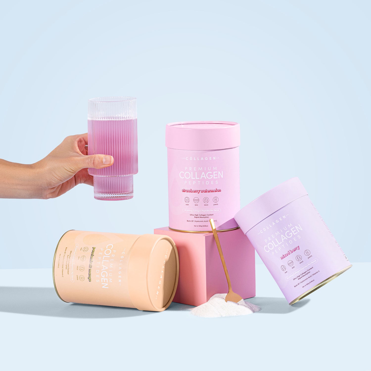 Hand holding glass of pink collagen drink, surrounded by pastel collagen containers and scoop on a light blue background.