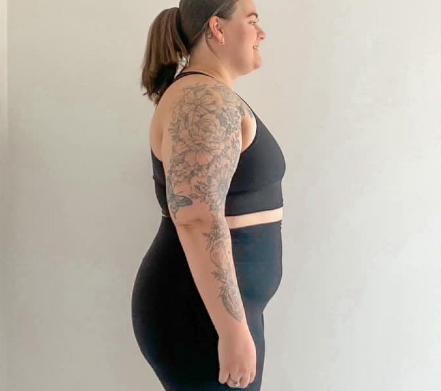 Profile of a woman in black workout attire, showcasing a floral arm tattoo, standing against a plain white background.