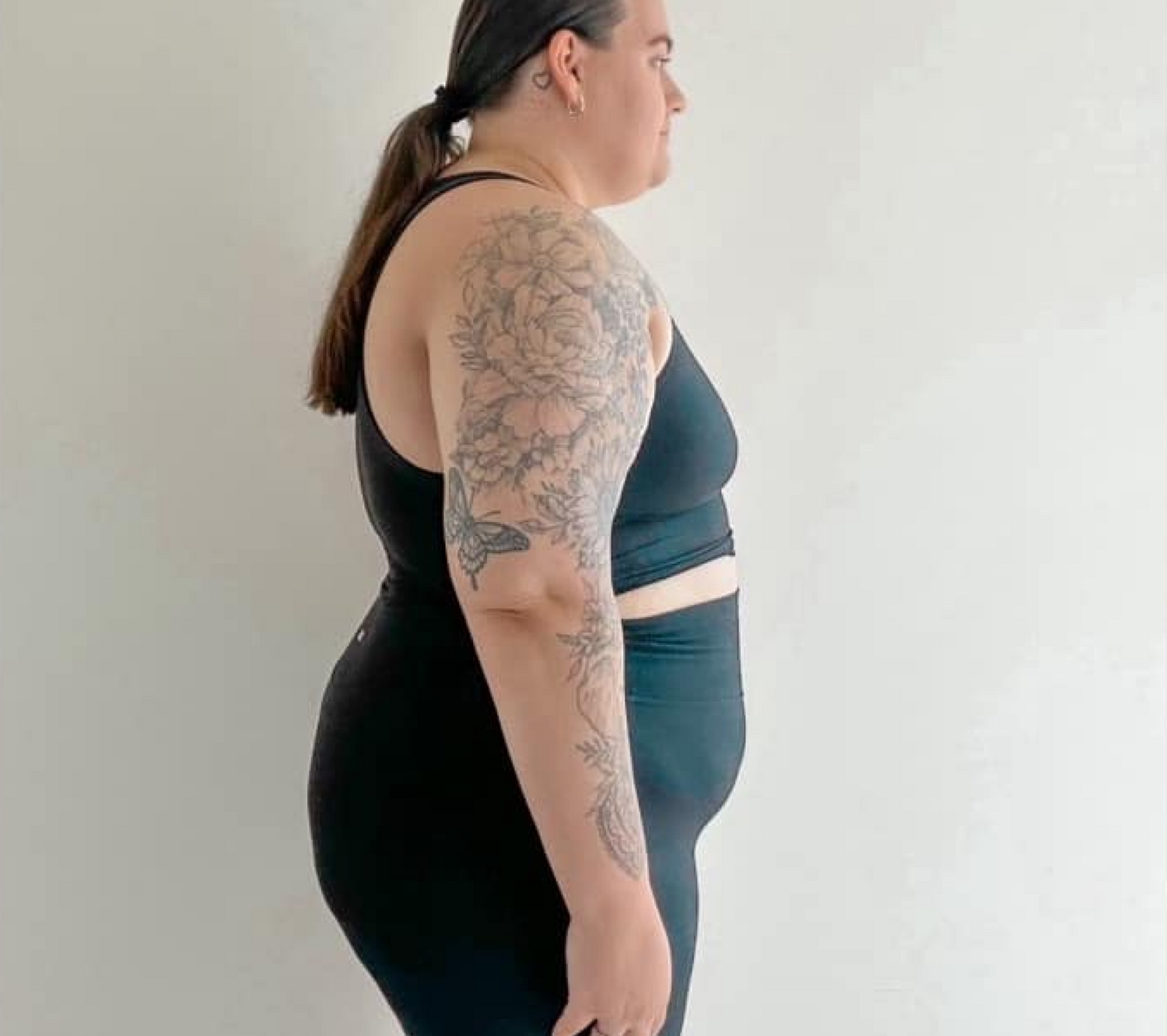 Person in black athletic wear with floral arm tattoos, standing in profile against a plain background.