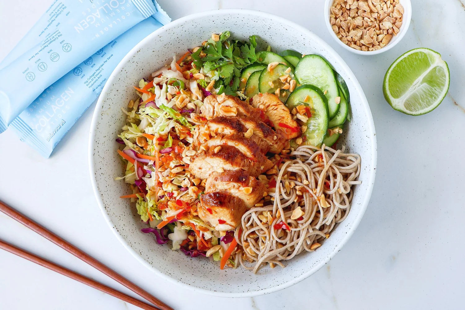 Nourishing bowl with grilled chicken, noodles, sliced cucumbers, and greens, accompanied by collagen packets, lime, and chopped nuts on a white surface.