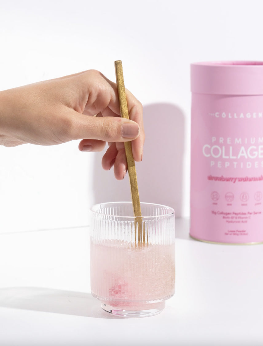 Hand stirring pink collagen drink in a clear glass, with a gold spoon beside a pink collagen container.