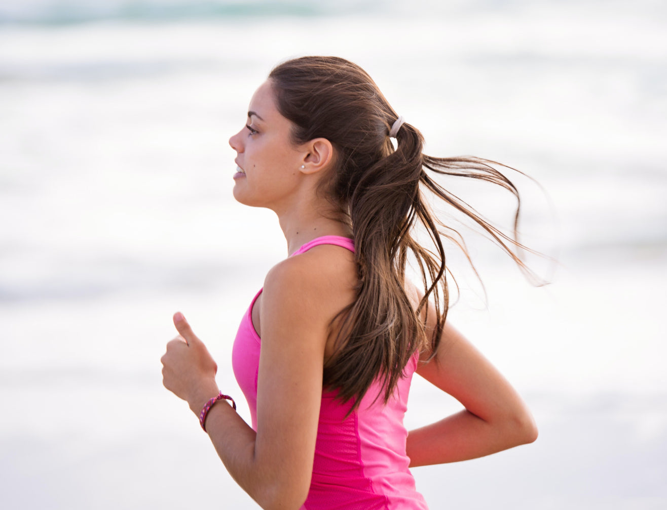 Woman jogging on beach in pink tank top, hair in ponytail, focused expression. Fitness, wellness, beauty, and healthy lifestyle.