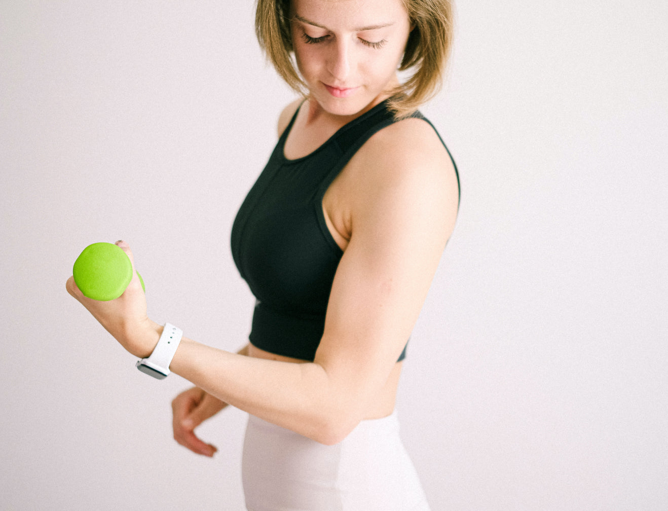 Woman in a black tank top exercises with a small green dumbbell. She wears a white watch and focuses on fitness and beauty.