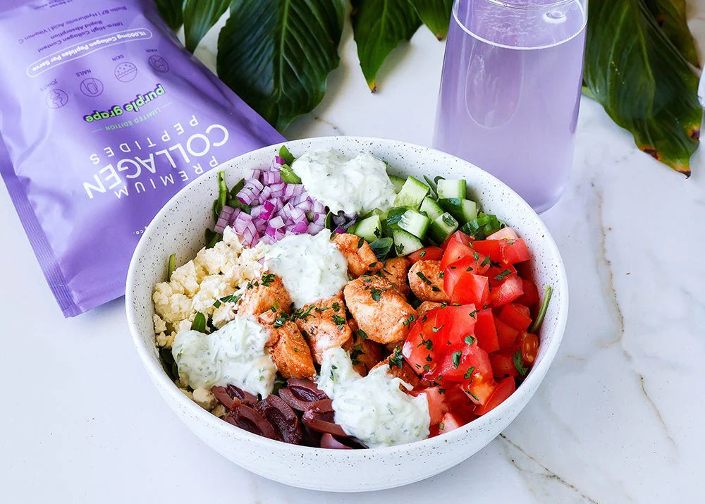 Colorful salad bowl with chicken, tomatoes, cucumbers, onions, and tzatziki sauce. Purple collagen supplement packet and glass nearby on white surface.