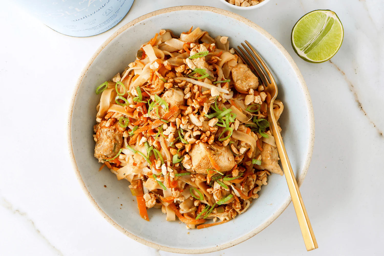A bowl of colorful pad thai with tofu, veggies, topped with peanuts; golden fork on side, lime wedge, and collagen container visible. Vegan, nutritious meal.