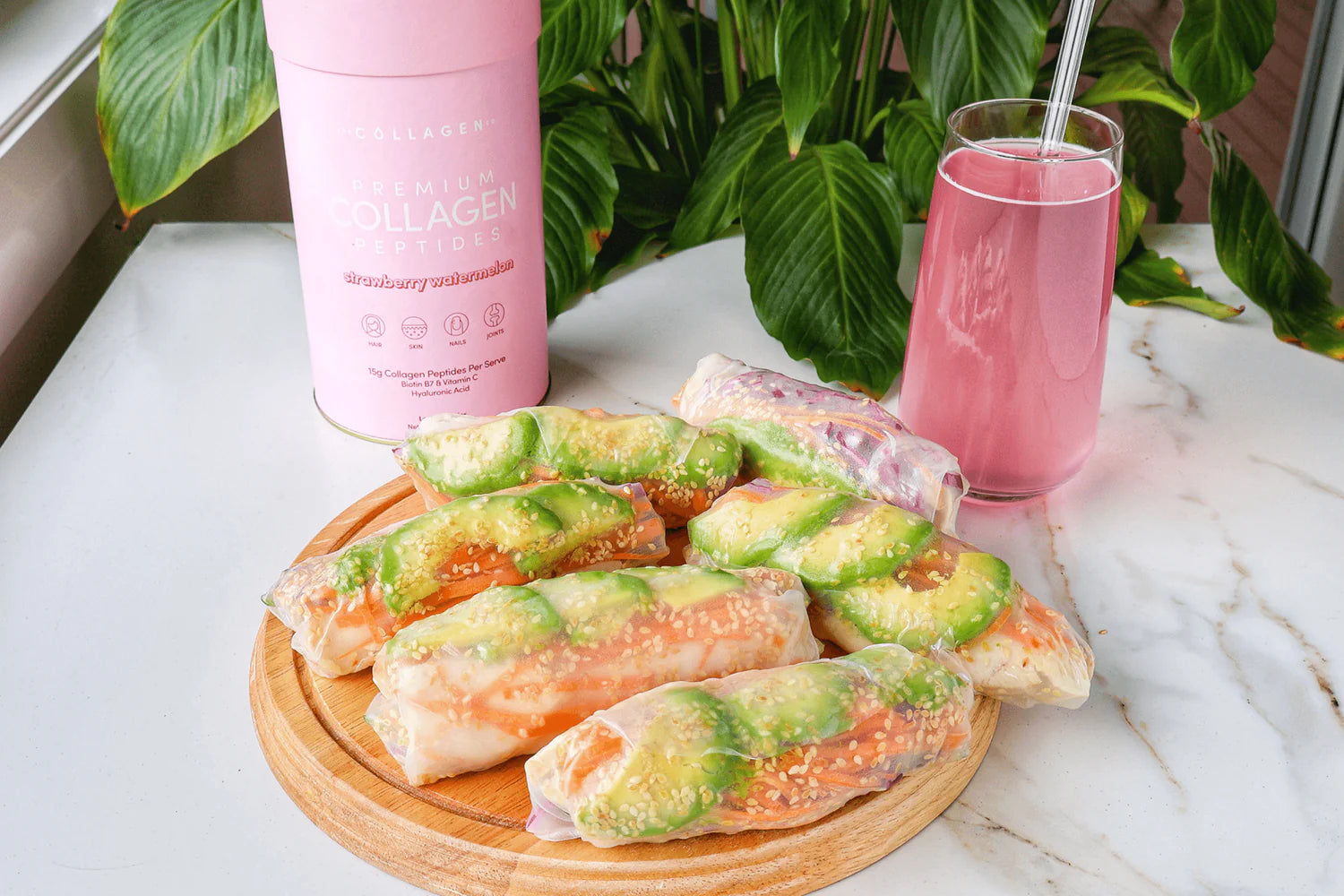 Fresh spring rolls with avocado and carrots on a wooden board, pink collagen drink in a glass, and collagen container in the background.