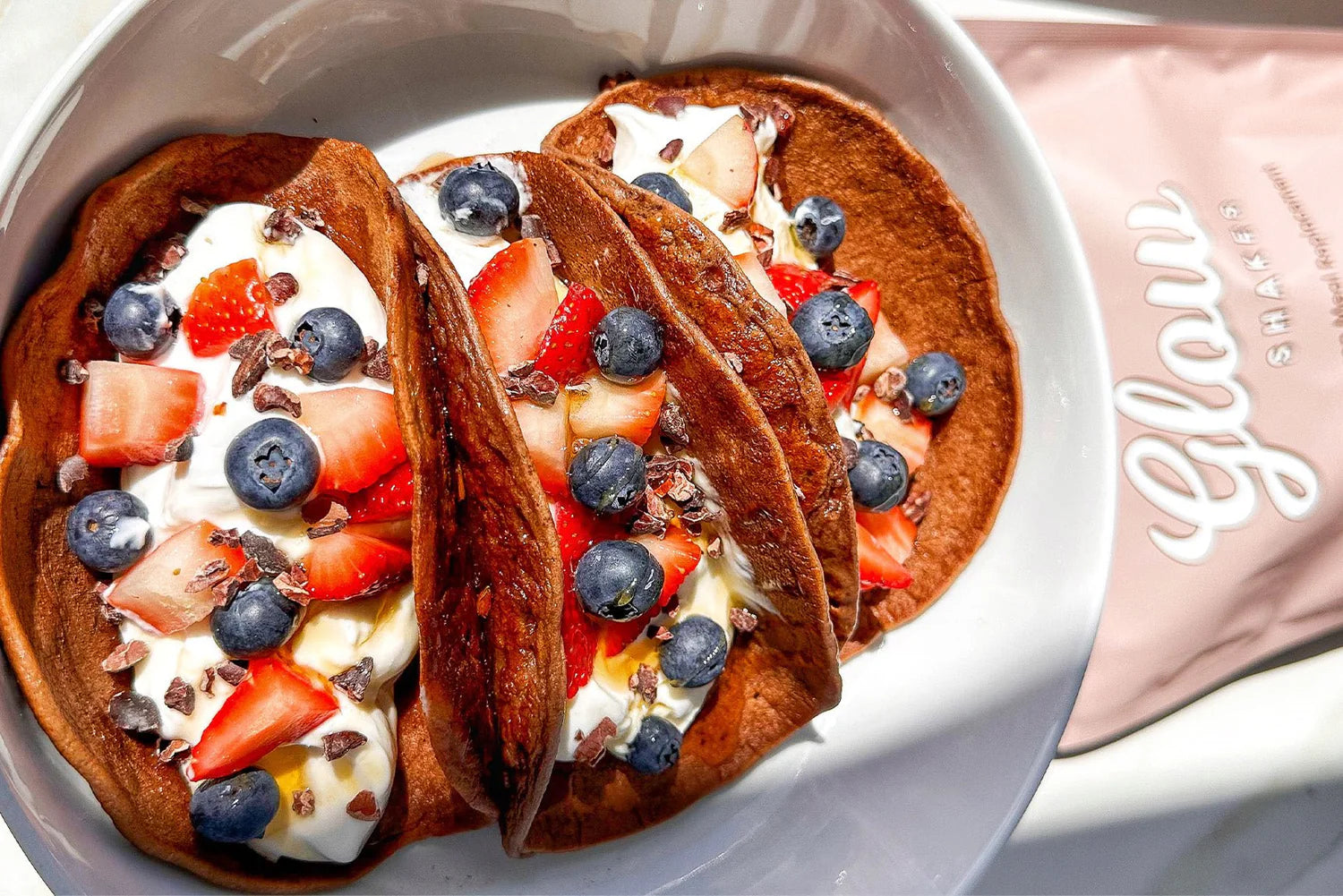 Chocolate pancakes with yogurt, strawberries, and blueberries in a bowl. "Glow" collagen supplement pack visible beside the bowl.