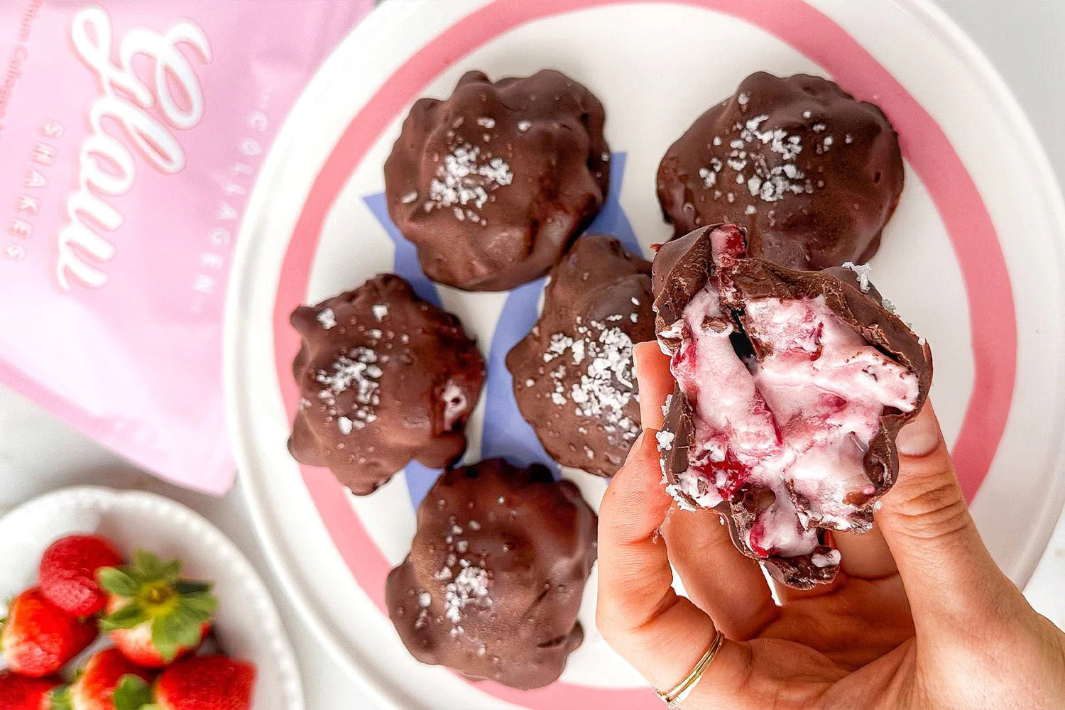 Chocolate-covered collagen bites with a creamy filling on a plate, a hand holding one bite, a packet of collagen in the background, and strawberries.