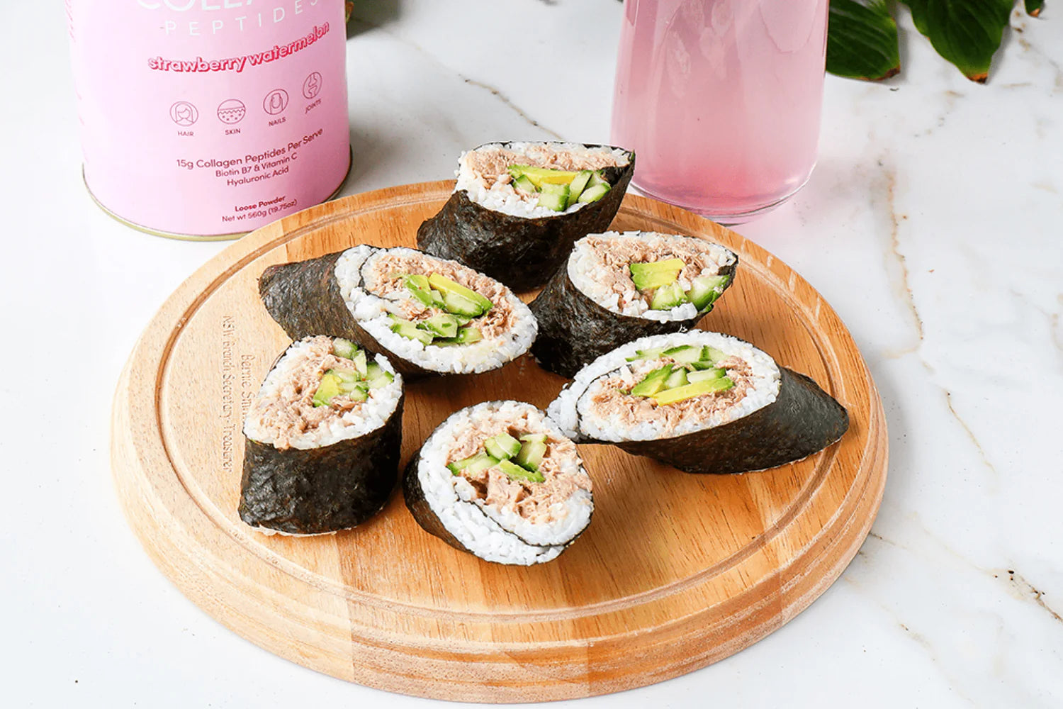 Sushi rolls with cucumber and tuna on a wooden board. A pink collagen supplement container and a glass with pink drink sit nearby.