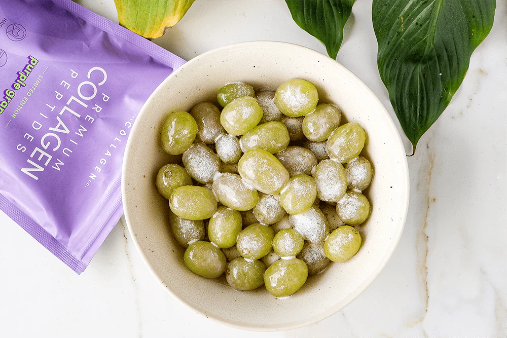 Bowl of frozen grapes dusted with white powder next to a purple collagen peptides packet on a marble surface with green leaves.