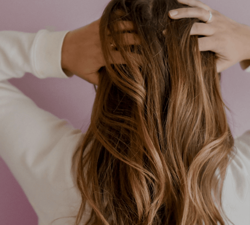 Woman with long, wavy hair running hands through it, wearing a white sweater against a pink background. Focus on hair health and beauty.