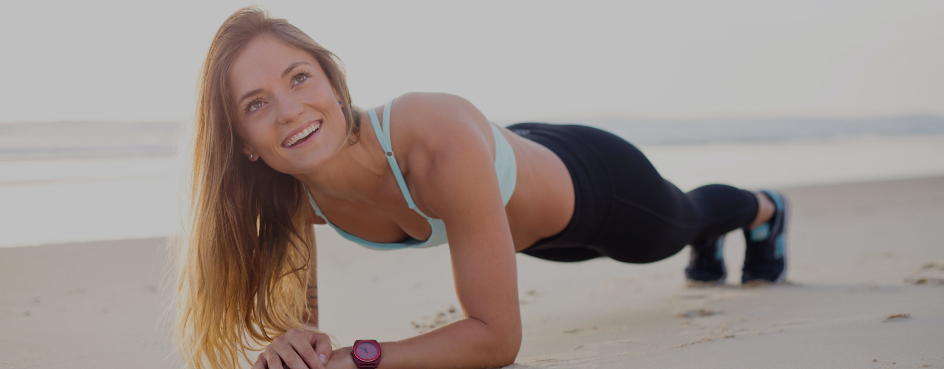 Smiling woman in a plank position on a beach, wearing a light blue sports bra and black leggings, with a watch.