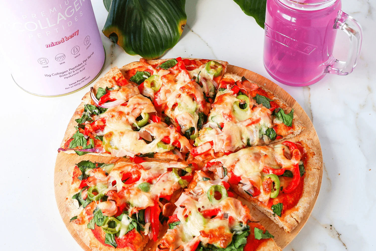 A veggie pizza sliced on a marble surface with a pink collagen supplement container and a mug of pink liquid beside it.