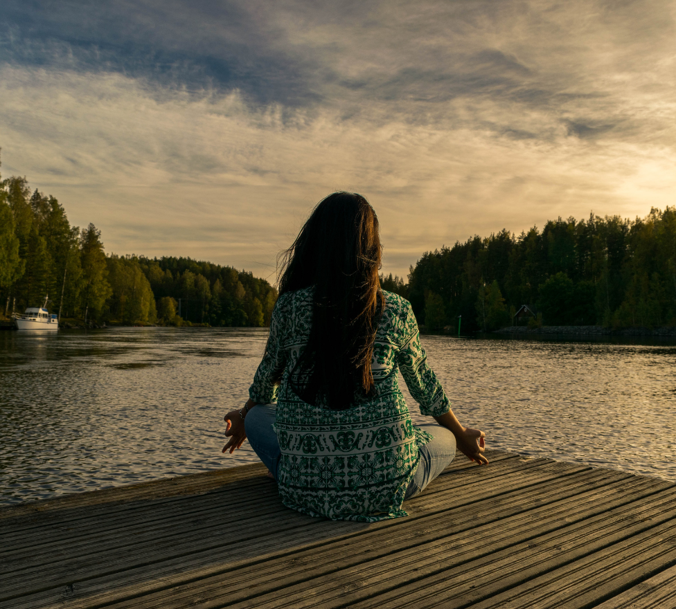 A woman in a patterned shirt meditates on a wooden pier by a tranquil lake at sunset, surrounded by lush trees.