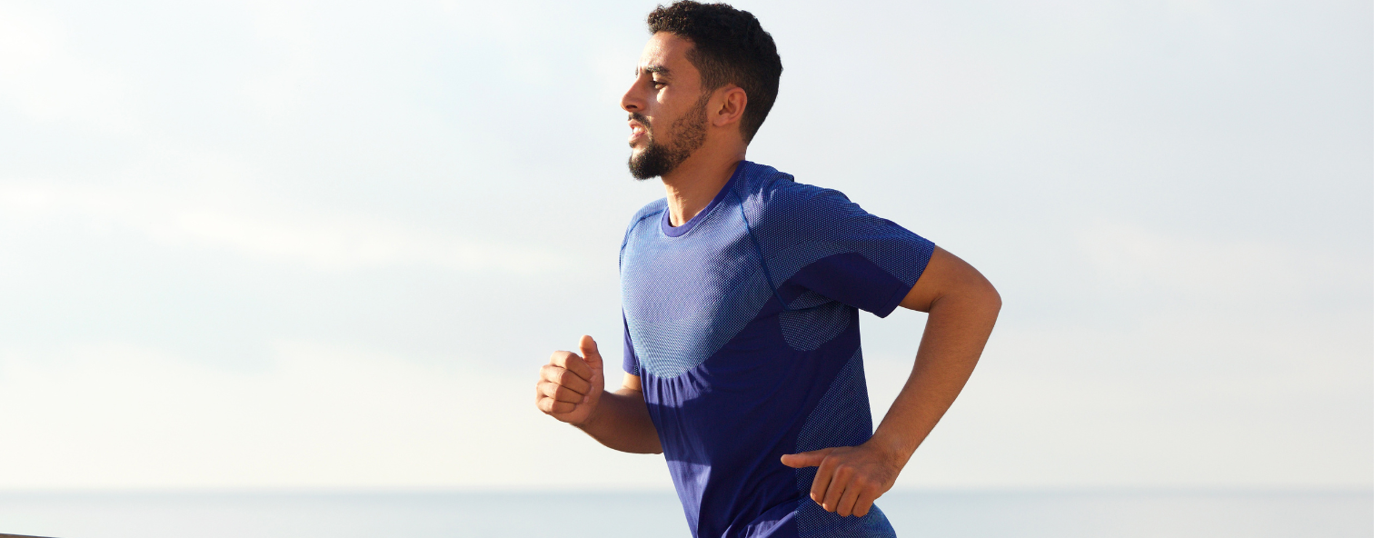 Man jogging near the beach in a blue shirt under a clear sky, focusing on fitness and wellness.