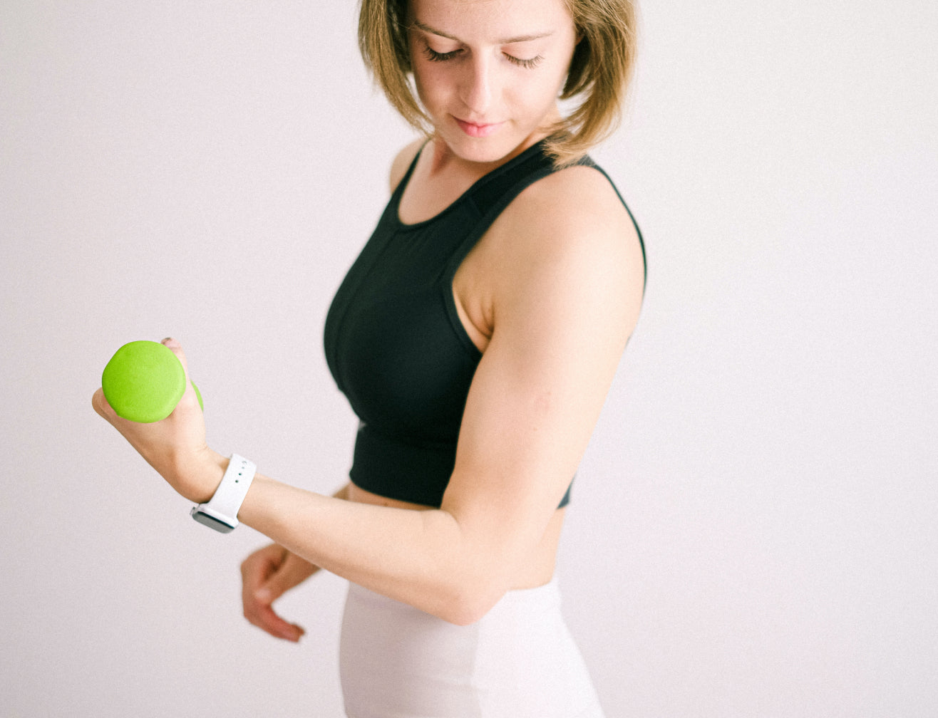 Woman in workout attire lifting green dumbbell, wearing a smartwatch. Focus on fitness, strength, and beauty.
