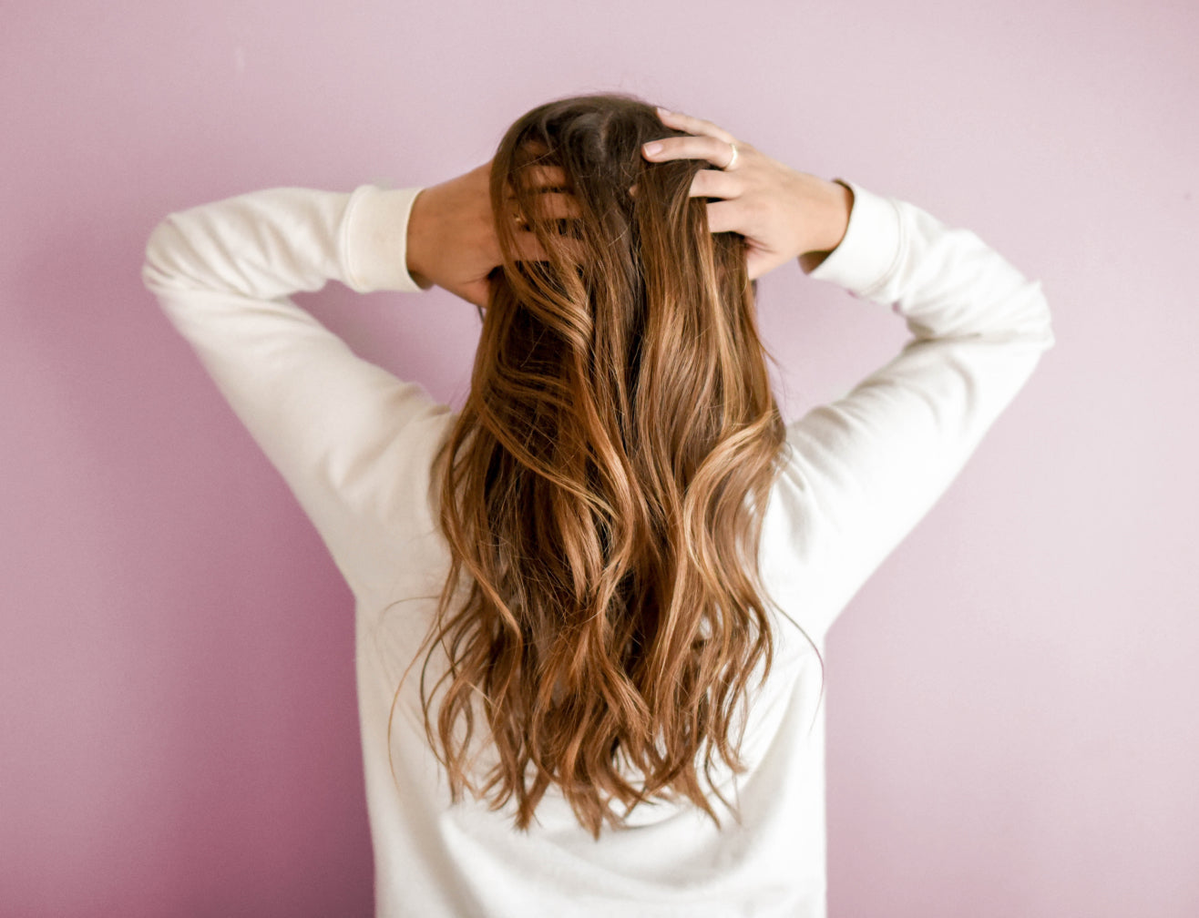 Woman with long, wavy hair facing a pink wall, hands in hair. Beauty and hair care emphasis.