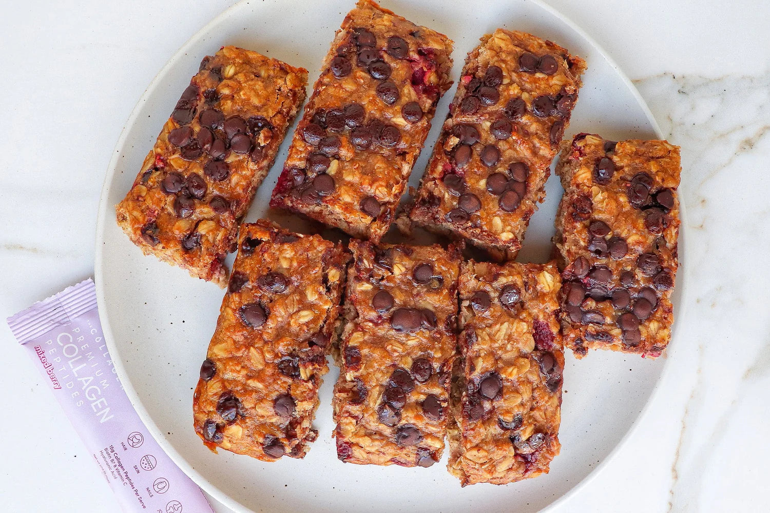 Collagen-infused granola bars with chocolate chips on a white plate, positioned on a marble surface; visible collagen packet beside.