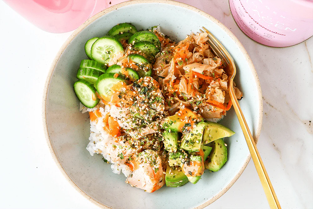 A bowl with rice, sliced cucumbers, avocado, kimchi, and salmon is topped with herbs and seeds. Pink collagen containers are partially visible.