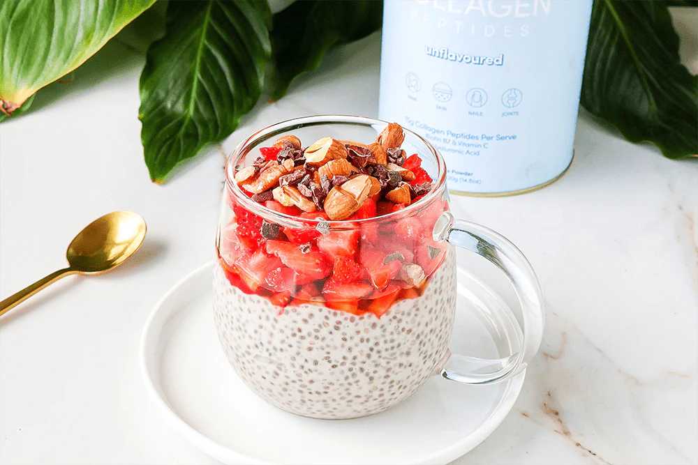 Glass mug filled with chia pudding, topped with fresh strawberries and nuts, placed on a white plate. Collagen supplement container in the background.